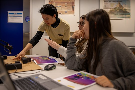 Frau zeigt zwei Schülerinnen etwas auf dem Tisch.